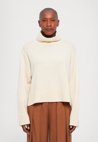 Osoba z ogoloną głową, ubrana w kremowy sweter z golfem i brązowe spodnie o wysokim stanie z plisami, stoi przodem.