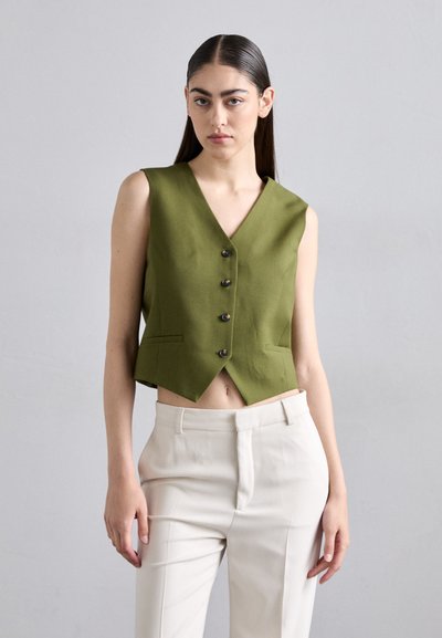 Groene cropped vest met een V-hals, mouwloos ontwerp en vijf zwarte knopen. Gecombineerd met lichtgekleurde broeken.