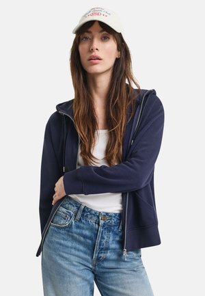 Veste à zip bleu marine avec un design ajusté, texture douce et capuche. Associée à un jean bleu clair et une casquette crème avec un texte.