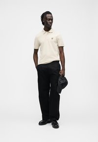 Polo beige con un piccolo logo giallo, abbinata a pantaloni cargo neri e scarpe nere; la persona tiene un berretto nero, in piedi davanti a uno sfondo uniforme.