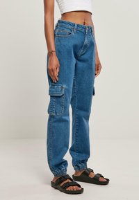 Pantalones cargo de mezclilla azul con puños elásticos, múltiples bolsillos laterales y un ajuste relajado. Llevados con sandalias negras sobre un fondo claro.