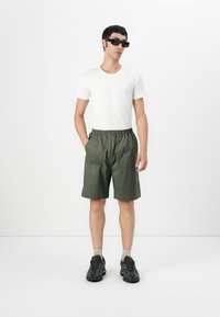 Grønne shorts med en avslappet passform, elastisk livlinje og to frontlommer, parret med en hvit t-skjorte og sorte joggesko.
