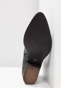 Zapato de tacón alto negro con parte superior de cuero texturizado y patrón en relieve. La parte inferior muestra una suela de madera plana y grabada con la talla 37.