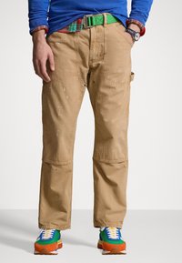 Bruna arbetsbyxor med synliga färgstänk, försedda med fickor och ett grönt bälte. Kombinerat med färgglada sneakers med blåa, gröna och orange detaljer.