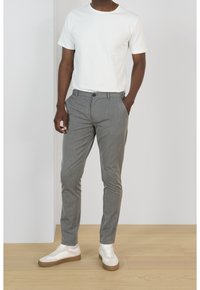 Bílý bavlněný tričko s krátkým rukávem spárované se šedými texturovanými slim-fit kalhotami a bílými teniskami s hnědými detaily. Stojí na dřevěné podlaze.