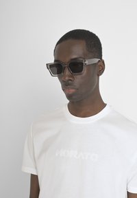 Gafas de sol negras de forma rectangular con un marco transparente, llevadas sobre una camiseta blanca lisa con un discreto texto en relieve que dice "MORATO".