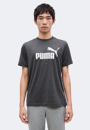 Junger Mann trägt ein dunkelgraues Puma T-Shirt mit Logo und hellgraue Hose und steht vor einem schlichten hellen Hintergrund.