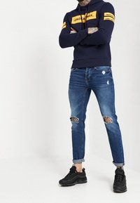 Sudadera de marina con detalles de logo amarillo, combinada con pantalones vaqueros azules desgastados que tienen desgarros en las rodillas y puños enrollados. Zapatillas negras.