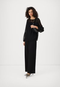 Blusa nera con maniche trasparenti a pois, abbinata a pantaloni neri a gamba larga e scarpe col tacco argento, mentre tiene una borsa nera con design ritagliato.