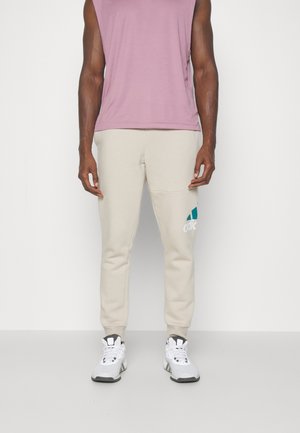 Homme portant un pantalon de survêtement beige Adidas avec un logo sur la cuisse, des baskets blanches et un haut sans manches rose poussière, debout devant un fond uni.