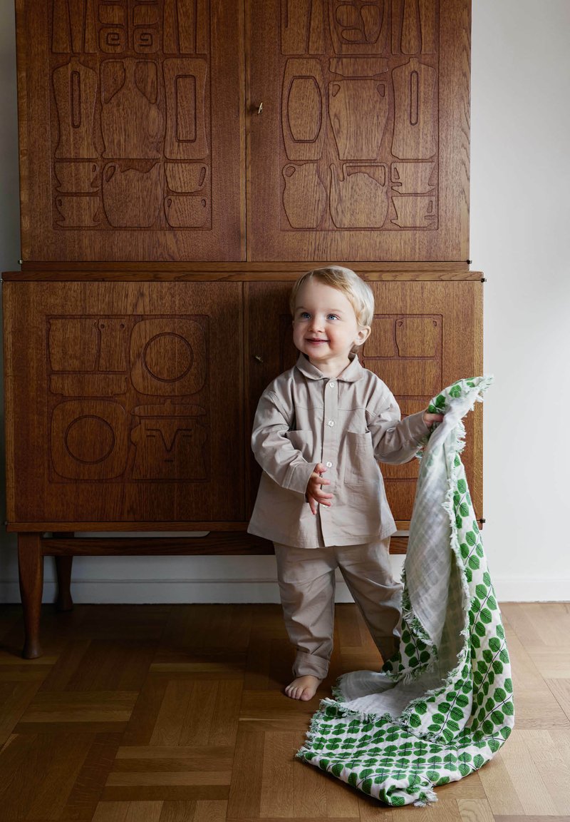 Lys beige bomulds pyjamas sæt på et barn, som holder et grønt og hvidt tæppe med bladmønster og fransede kanter, foran et udskåret træskab.