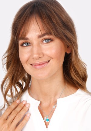 Collier en argent avec un pendentif en forme d'œil orné d'une pierre turquoise, accentué par de petits détails métalliques. Modèle portant une blouse blanche.