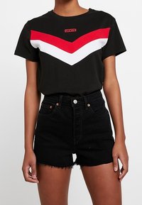 Camiseta negra con rayas en forma de chevrón rojas y blancas en el pecho, con el logo de Levi's. Un par de pantalones cortos de mezclilla negros de tiro alto con dobladillo deshilachado.
