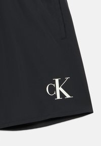 Μαύρο σορτς με λεία υφή, που διαθέτει το εμφανές λευκό λογότυπο "cK" στο κάτω αριστερό μέρος και πλαϊνές τσέπες. Απλός σχεδιασμός, χωρίς σχέδια.