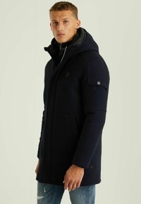 Tamsiai mėlynas gobtuvu paltas su užtrauktuku, šoninėmis kišenėmis ir išklota vidine dalimi. Yra logotipo detalė ir tekstūruota medžiaga.