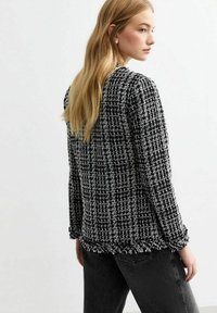 Zwart-witte tweedjas met een gemêleerd patroon, voorzien van rafelige randen en lange mouwen. In combinatie met donkergrijze jeans.