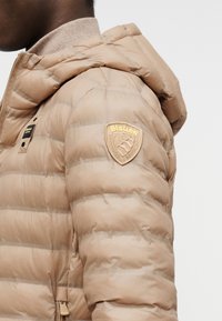 Πλάγια όψη προσώπου που φοράει μπεζ μπουφάν με κουκούλα και το λογότυπο της Blauer στο άνω μέρος του χεριού, με ορατή τσέπη με φερμουάρ κοντά στη μέση.