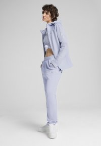 Ensemble de sport violet clair constitué d'une veste à capuche et de pantalons de jogging, avec un crop top noué. Des baskets blanches complètent le look.