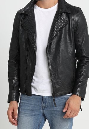 Hombre con chaqueta de cuero negra con solapas acolchadas sobre una camiseta blanca y jeans azules, de pie contra un fondo liso.