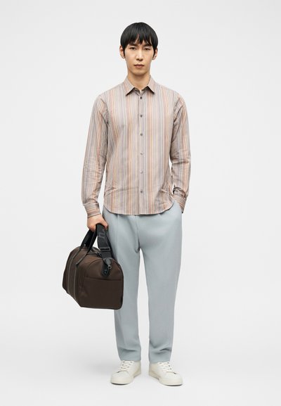 Άνδρας που φοράει ριγέ πουκάμισο με κουμπιά, ανοιχτό γκρι παντελόνι, λευκά αθλητικά παπούτσια, κρατώντας καφέ τσάντα ταξιδιού σε ουδέτερο φόντο.