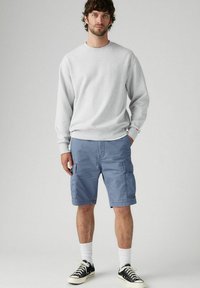 Man wearing light gray sweatshirt, blue cargo shorts, white socks, and black low-top sneakers standing against plain gray background.