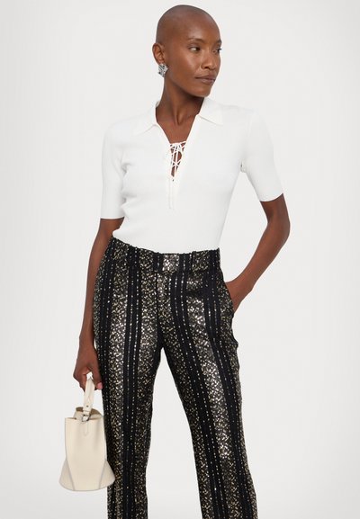 White ribbed knit top with a lace-up neckline paired with black sequined pants featuring gold stripes. Carrying a small white handbag.