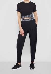 Czarna krótka koszulka i czarne dresy z szerokim elastycznym pasem, na którym widnieje logo "Calvin Klein", w połączeniu z czarnymi butami sportowymi.