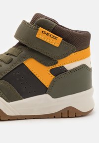 Zapatilla alta para niños en verde, negro y naranja con correa de Velcro etiquetada como "GEOX", cuello acolchado marrón y suela blanca con relieve marrón.