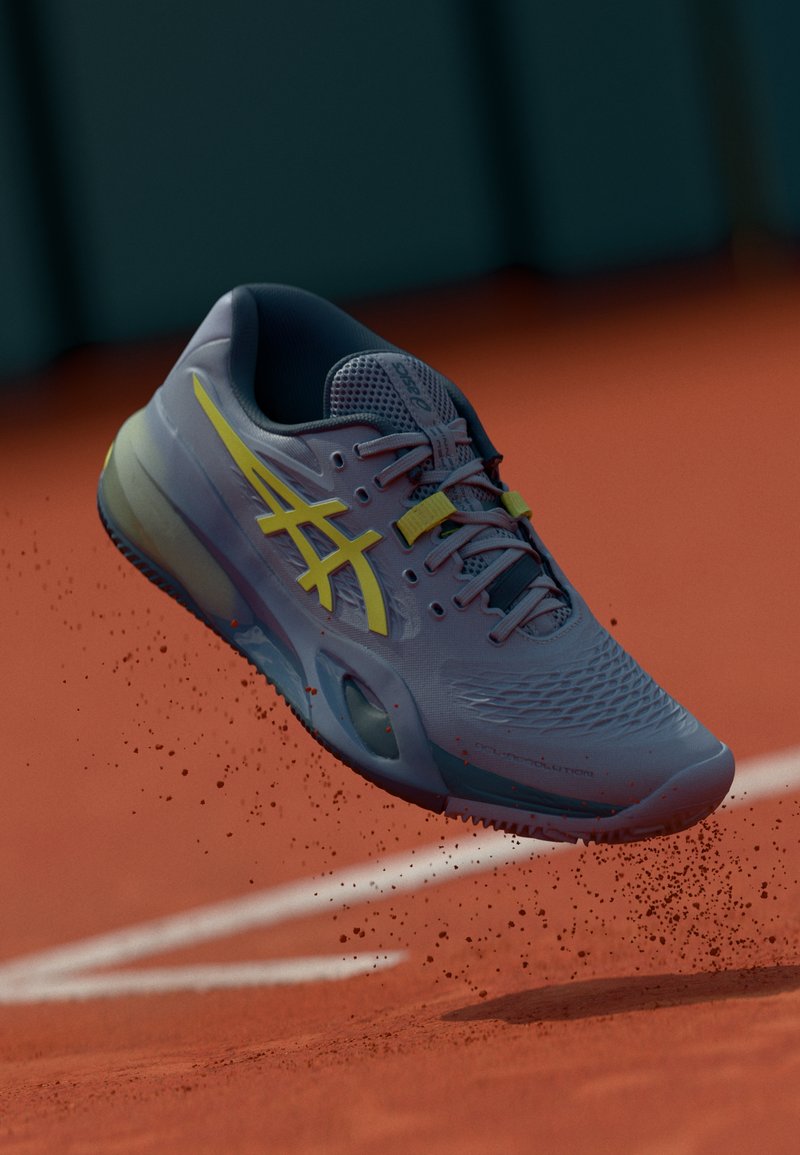 Une chaussure de course bleue et jaune qui soulève de la poussière d'argile, capturée en plein vol au-dessus d'un court de tennis extérieur rouge, près de la ligne blanche de fond.