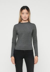 Giovane donna con capelli lunghi e lisci, che indossa un top aderente grigio scuro a coste con maniche lunghe e pantaloni neri, in piedi davanti a uno sfondo semplice.