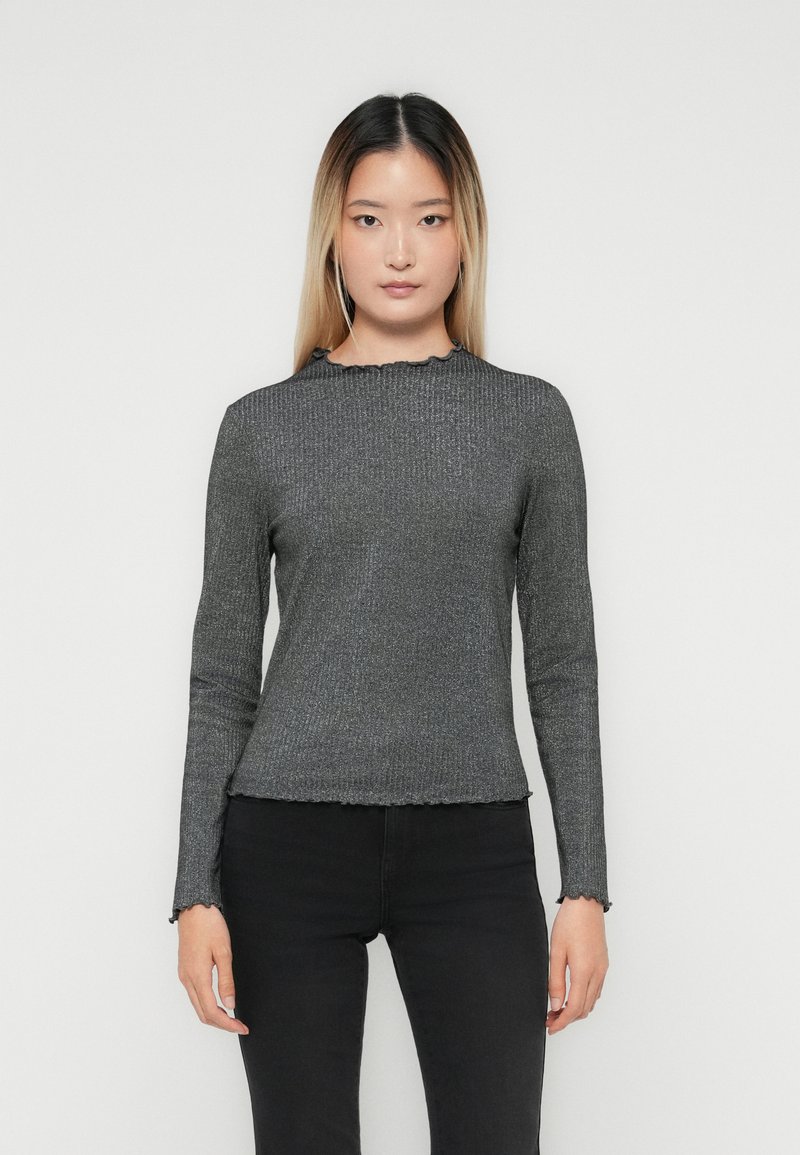 Giovane donna con capelli lunghi e lisci, che indossa un top aderente grigio scuro a coste con maniche lunghe e pantaloni neri, in piedi davanti a uno sfondo semplice.