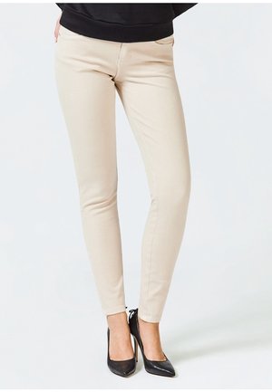 Beige slim-fit broek gedragen met zwarte puntige hoge hakken, getoond van taille tot voeten tegen een effen achtergrond.