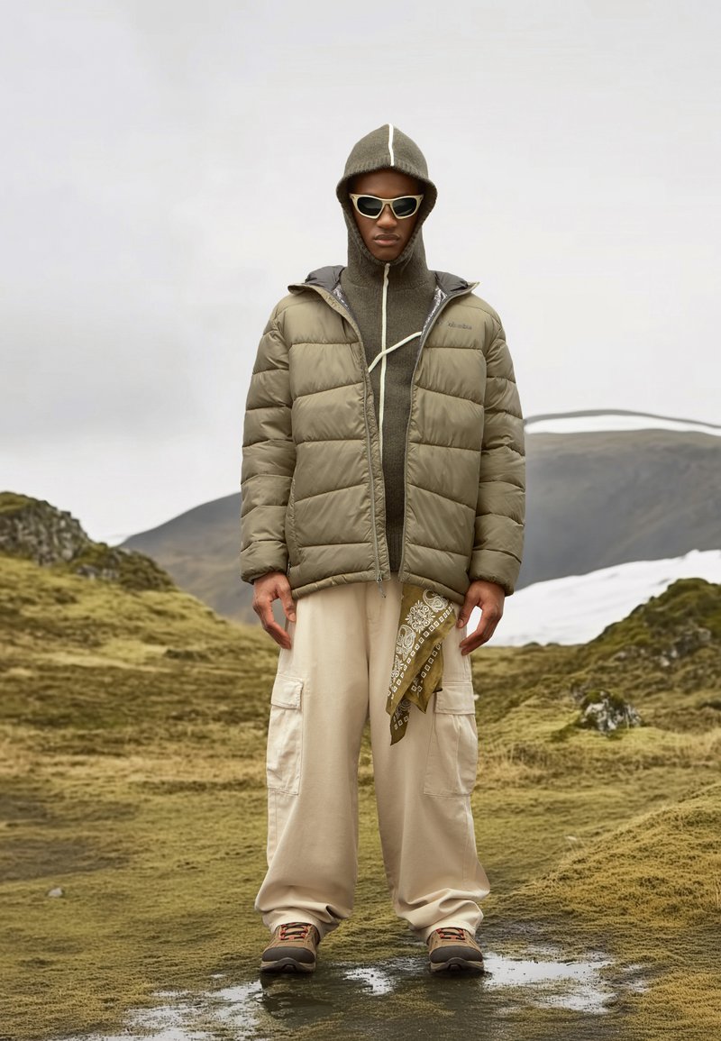 Chaqueta acolchada color oliva con capucha y cremallera en contraste, pantalones cargo beige, zapatos marrones y un pañuelo con estampado atado a la cintura.