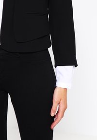 Musta istuva takki, jossa on sileä pinta, yhdistettynä mustiin housuihin ja näkyvissä valkoiseen paitaan kuuluvaan hihaan. Takissa on rakenteelliset kannukset.