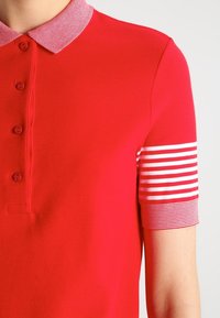 Červená polokošile s texturovanou tkaninou, která má kontrastní proužkovaný límec a proužkovaný akcent na rukávech. Detail s knoflíkovou légou na čtyři knoflíky.