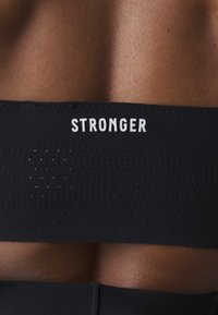 Melnā sporta krūšturis ar baltu uzrakstu "STRONGER". Iezīmēts ar perforētu tekstūru, lai nodrošinātu elpojamību, un gludu, pieguļošu dizainu.