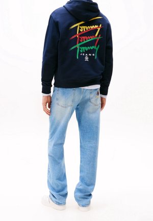 Person trägt marineblauen Hoodie mit buntem "Tommy Jeans"-Logo auf dem Rücken, hellblaue Jeans mit Aufnäher auf der Tasche und weiße Turnschuhe, steht mit dem Rücken zur Kamera.