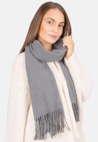 Femme aux cheveux bruns lisses portant une écharpe grise à franges et un pull en tricot crème, tenant l'écharpe près de son cou devant un fond blanc.