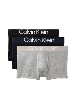 Homme musclé debout pieds nus portant un boxer noir Calvin Klein sur un fond gris uni.