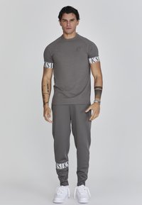 Grijs sportoutfit bestaande uit een t-shirt met korte mouwen met logo op de borst en gestreepte boorden, gecombineerd met taps toelopende joggers; witte sneakers.