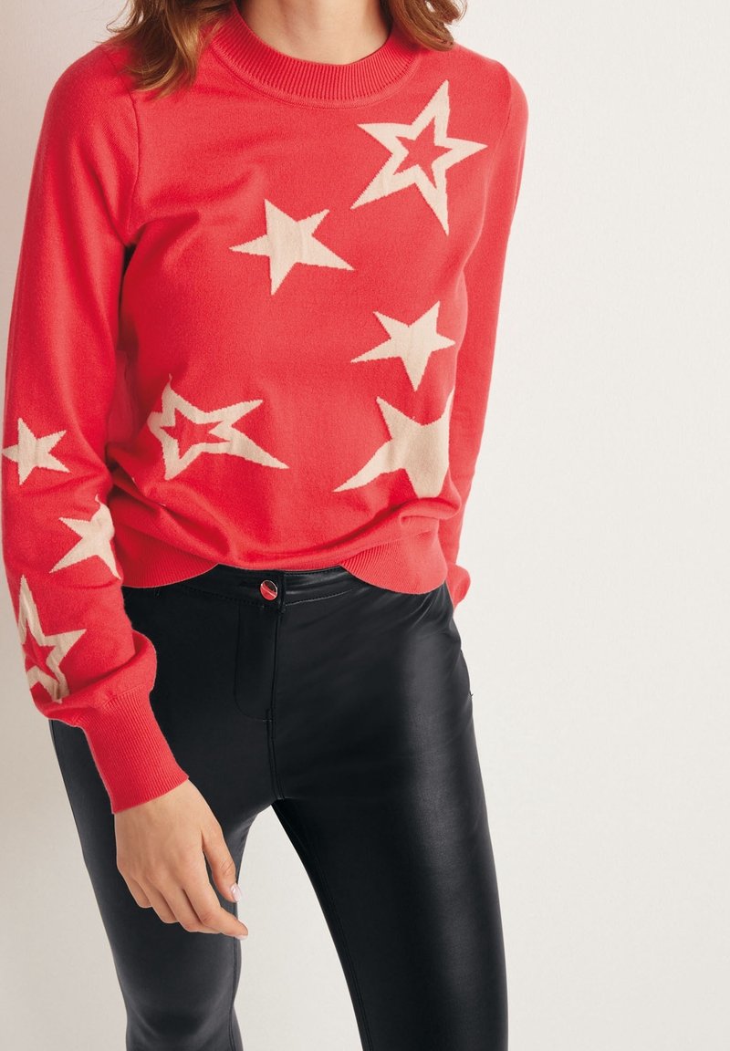 Roter Pullover mit cremefarbenen Sternmustern, Rundhals-Ausschnitt und gerippten Bündchen und Saum, getragen mit schwarzen Lederhosen mit einem roten Knopf.