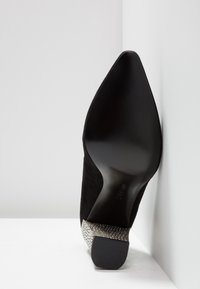 Soulier à talon haut en suède noir avec un bout pointu et un talon métallique texturé. Semelle lisse avec un détail de marque discret.