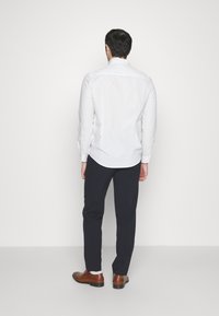 Chemise blanche à manches longues, design ajusté, associée à un pantalon de costume bleu marine, chaussures en cuir marron, portée par un modèle vu de dos.