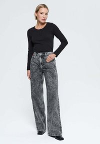 Femme se tenant contre un fond blanc, portant un haut noir à manches longues, un jean large taille haute en délavé acide et des chaussures noires à bout carré.