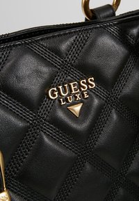 Black quilted handbag made of synthetic leather with a gold "GUESS LUXE" logo and triangular accent. Features textured surface and gold hardware.