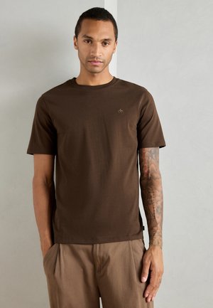 Braunes Baumwoll-T-Shirt mit kurzen Ärmeln, Rundhalsausschnitt und einem kleinen Logo auf der linken Brust. Glatte Textur und lockere Passform, kombiniert mit beigen Hosen.