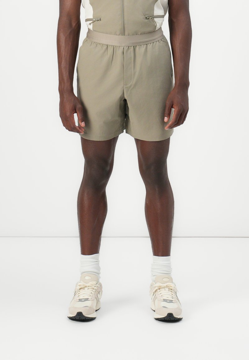 Shorts pour hommes de couleur vert olive avec une taille élastique, dotés d'une fermeture éclair à l'avant et de poches latérales, associés à des baskets et des chaussettes de couleur claire.