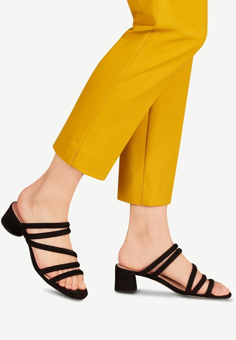 Pieds portant des sandales à talons noires à brides, associées à un pantalon court jaune moutarde sur un fond clair.