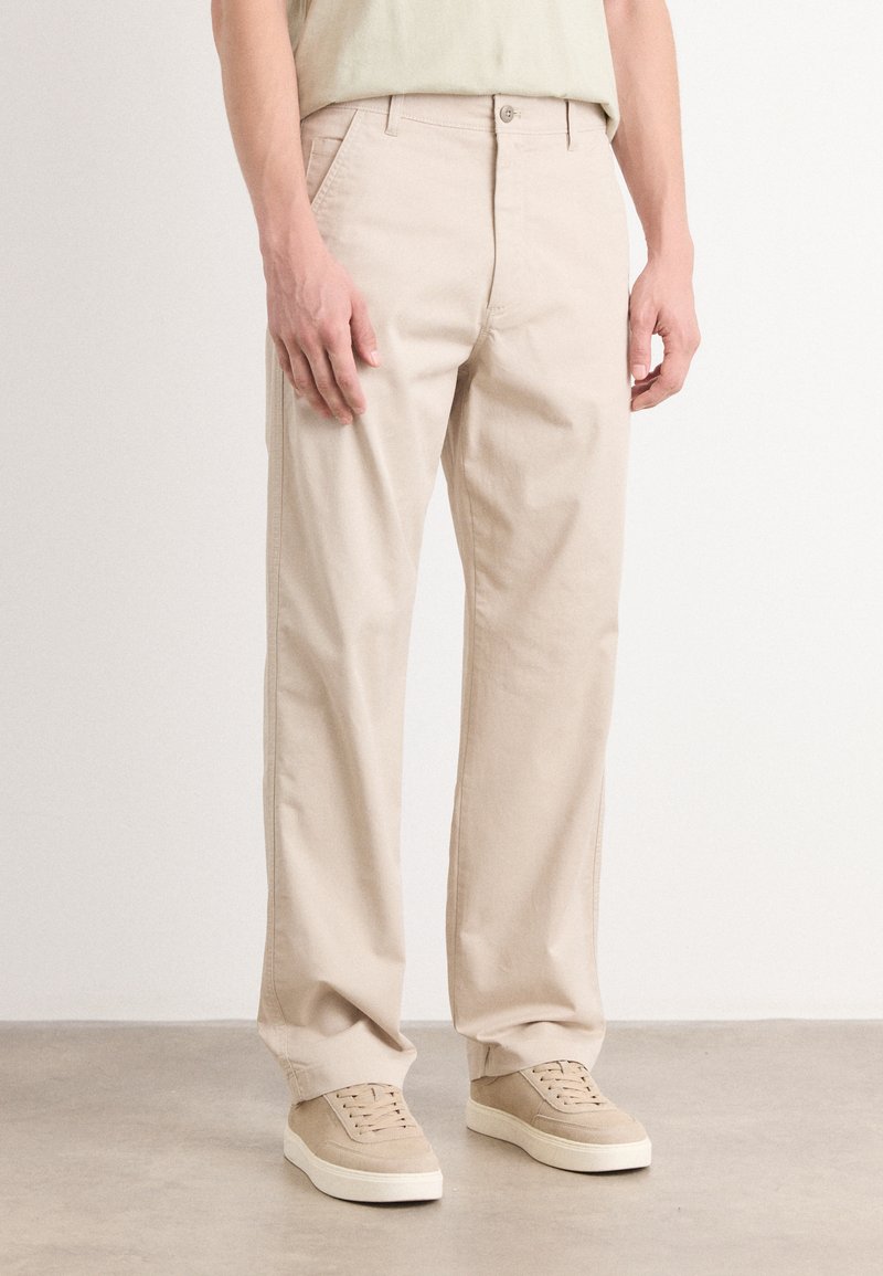 Beige bomullsbukser med rett snitt, med sidelommer og glidelås, parret med beige joggesko på en jevn overflate.