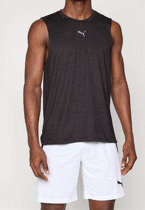 Uomo che indossa una canottiera sportiva nera senza maniche Puma e pantaloncini bianchi Puma, in piedi contro uno sfondo chiaro uniforme.